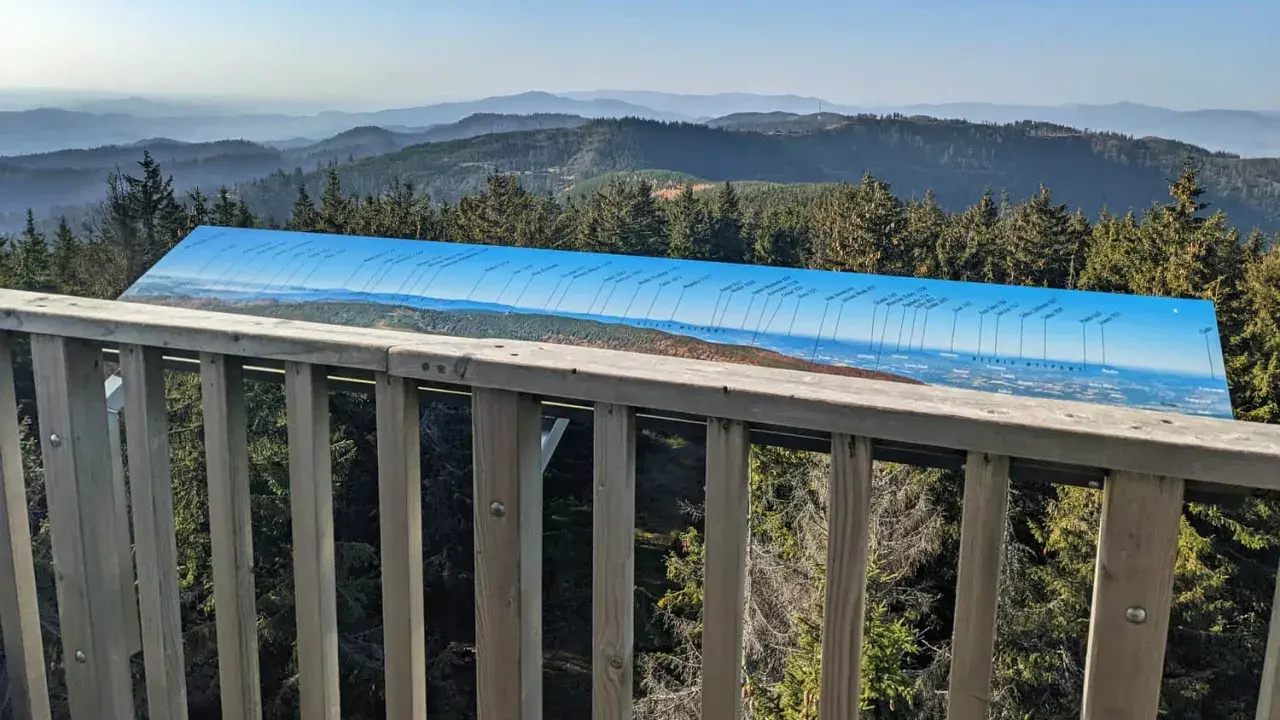 Widok z wieży widokowej na Beskid Sądecki. Na pierwszym planie drewniana balustrada, dalej panorama gór i lasów z tablicą informacyjną o szczytach.