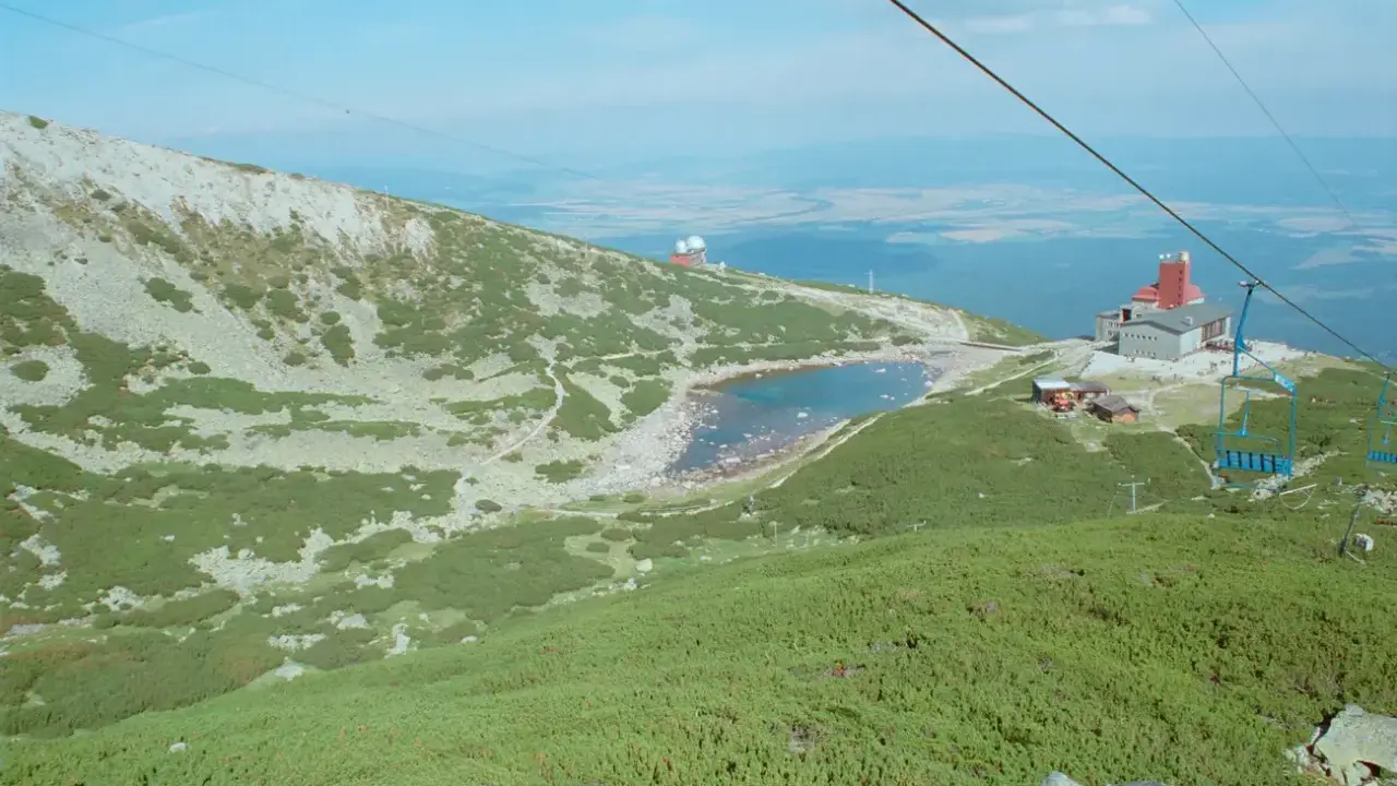Widok na skaliste pleso otoczone zielenią i kamieniami. W oddali budynek z czerwoną wieżą i kolejka linowa.