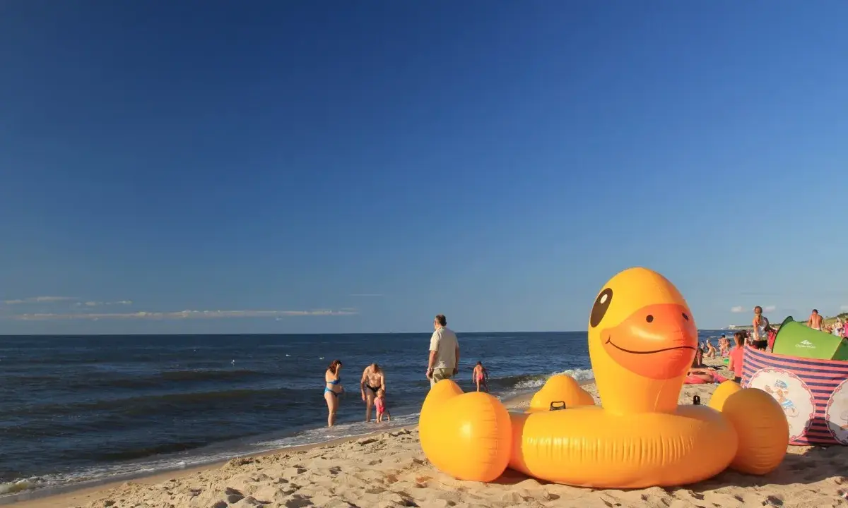 Słoneczna plaża w Bobolinie, dmuchana kaczka i inne atrakcje dla dzieci. Rodziny kąpią się w morzu.