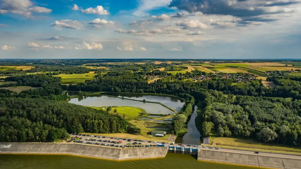 Malowniczy krajobraz z jeziorem Chańcza, zaporą, lasami i polami uprawnymi pod błękitnym, chmurowym niebem.