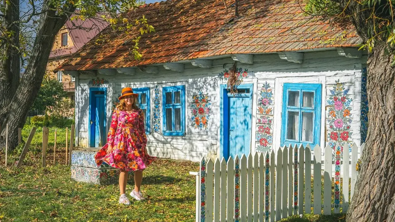 Kobieta w kolorowej sukience i kapeluszu przed malowanymi domami w Zalipiu. Wiosna w pełni zdobi chaty.