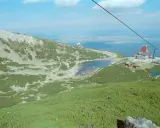 Widok na skaliste pleso otoczone zielenią i kamieniami. W oddali budynek z czerwoną wieżą i kolejka linowa.