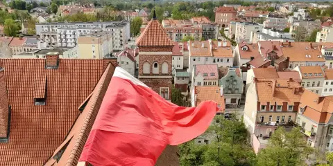 Widok na miasto z polską flagą na pierwszym planie. W tle widać zabytkowe kamienice i wieżę kościelną, co sugeruje ciekawe lidzbark warmiński atrakcje.
