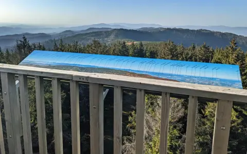 Widok z wieży widokowej na Beskid Sądecki. Na pierwszym planie drewniana balustrada, dalej panorama gór i lasów z tablicą informacyjną o szczytach.
