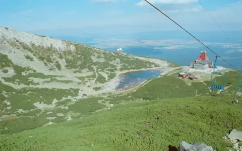 Widok na skaliste pleso otoczone zielenią i kamieniami. W oddali budynek z czerwoną wieżą i kolejka linowa.