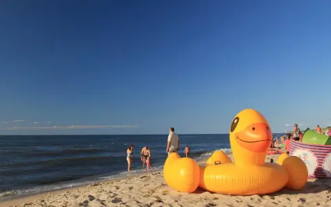 Słoneczna plaża w Bobolinie, dmuchana kaczka i inne atrakcje dla dzieci. Rodziny kąpią się w morzu.