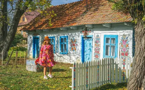 Kobieta w kolorowej sukience i kapeluszu przed malowanymi domami w Zalipiu. Wiosna w pełni zdobi chaty.