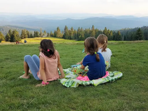 Trzy dziewczynki odpoczywają na kocu, podziwiając malowniczy widok gór i lasów. Może to właśnie szlak rysianka?