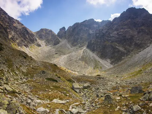 Kamienista dolina w Tatrach Wysokich, gdzie wije się szlak. W tle skaliste szczyty i błękitne niebo z chmurami.