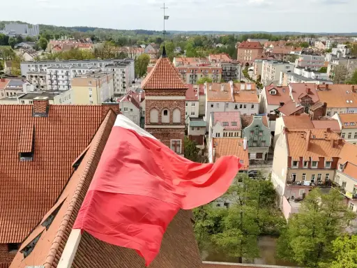 Widok na miasto z polską flagą na pierwszym planie. W tle widać zabytkowe kamienice i wieżę kościelną, co sugeruje ciekawe lidzbark warmiński atrakcje.