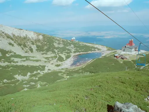 Widok na skaliste pleso otoczone zielenią i kamieniami. W oddali budynek z czerwoną wieżą i kolejka linowa.