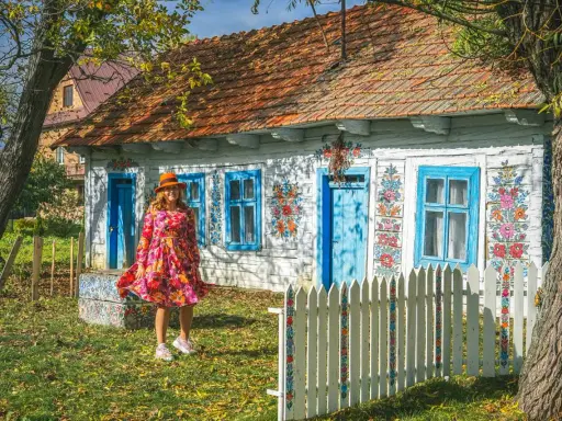 Kobieta w kolorowej sukience i kapeluszu przed malowanymi domami w Zalipiu. Wiosna w pełni zdobi chaty.
