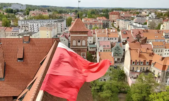 Widok na miasto z polską flagą na pierwszym planie. W tle widać zabytkowe kamienice i wieżę kościelną, co sugeruje ciekawe lidzbark warmiński atrakcje.