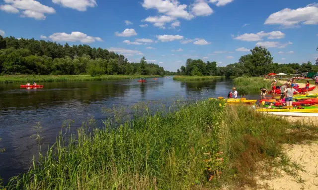 Spływ kajakowy w Załęczańskim Parku Krajobrazowym. Ludzie przygotowują się do wodnej przygody na piaszczystej plaży nad rzeką.