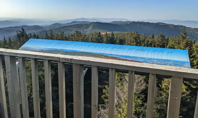 Widok z wieży widokowej na Beskid Sądecki. Na pierwszym planie drewniana balustrada, dalej panorama gór i lasów z tablicą informacyjną o szczytach.
