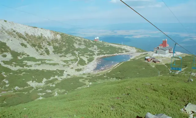 Widok na skaliste pleso otoczone zielenią i kamieniami. W oddali budynek z czerwoną wieżą i kolejka linowa.