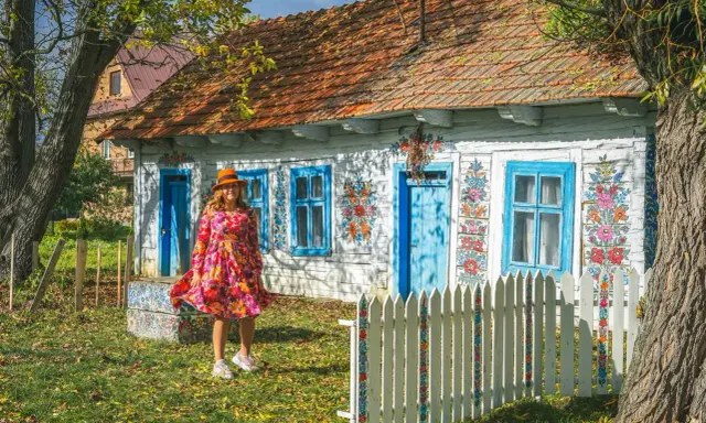 Kobieta w kolorowej sukience i kapeluszu przed malowanymi domami w Zalipiu. Wiosna w pełni zdobi chaty.