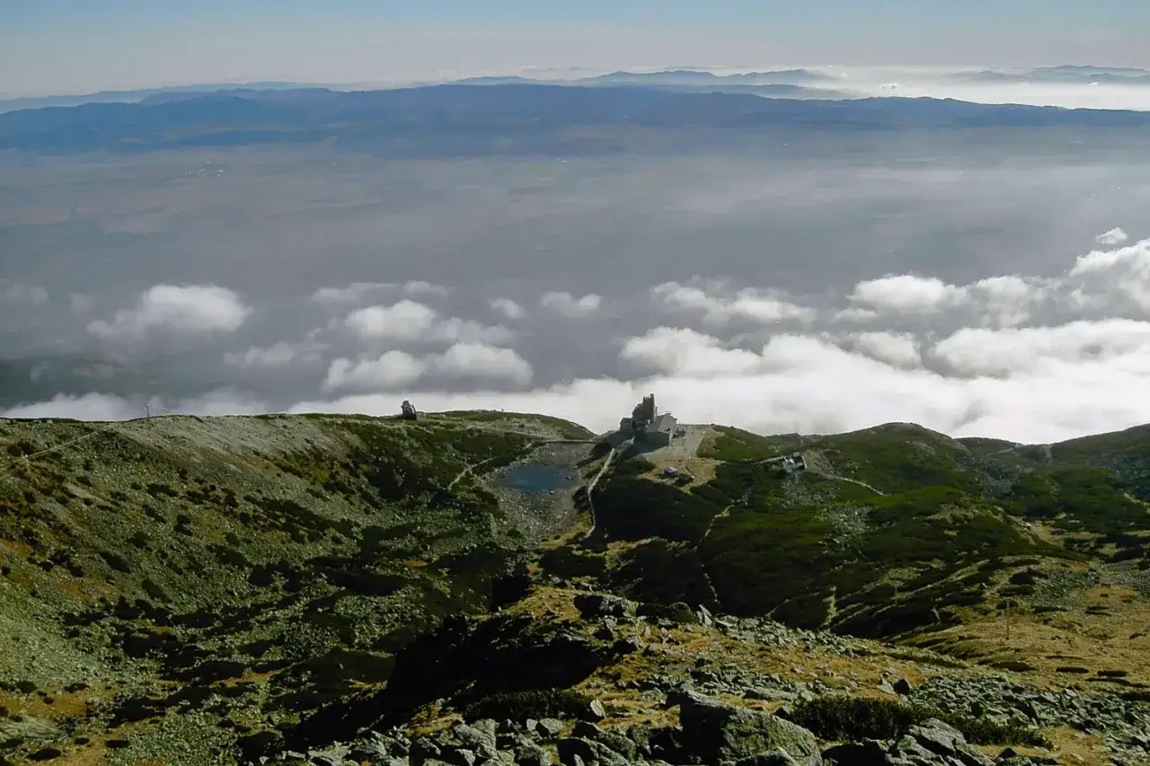 Widok na skaliste pleso otoczone g&oacute;rami, z budynkami obserwatorium na szczycie.