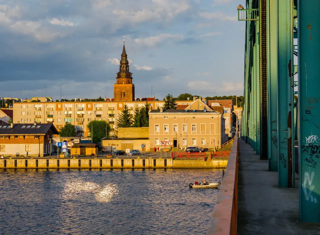 Zach&oacute;d słońca nad Gryfinem. Widok na port, wieżę kościelną i most. Woda odbija światło, a na niej płynie ł&oacute;dka. To jedne z wielu gryfino atrakcji.
