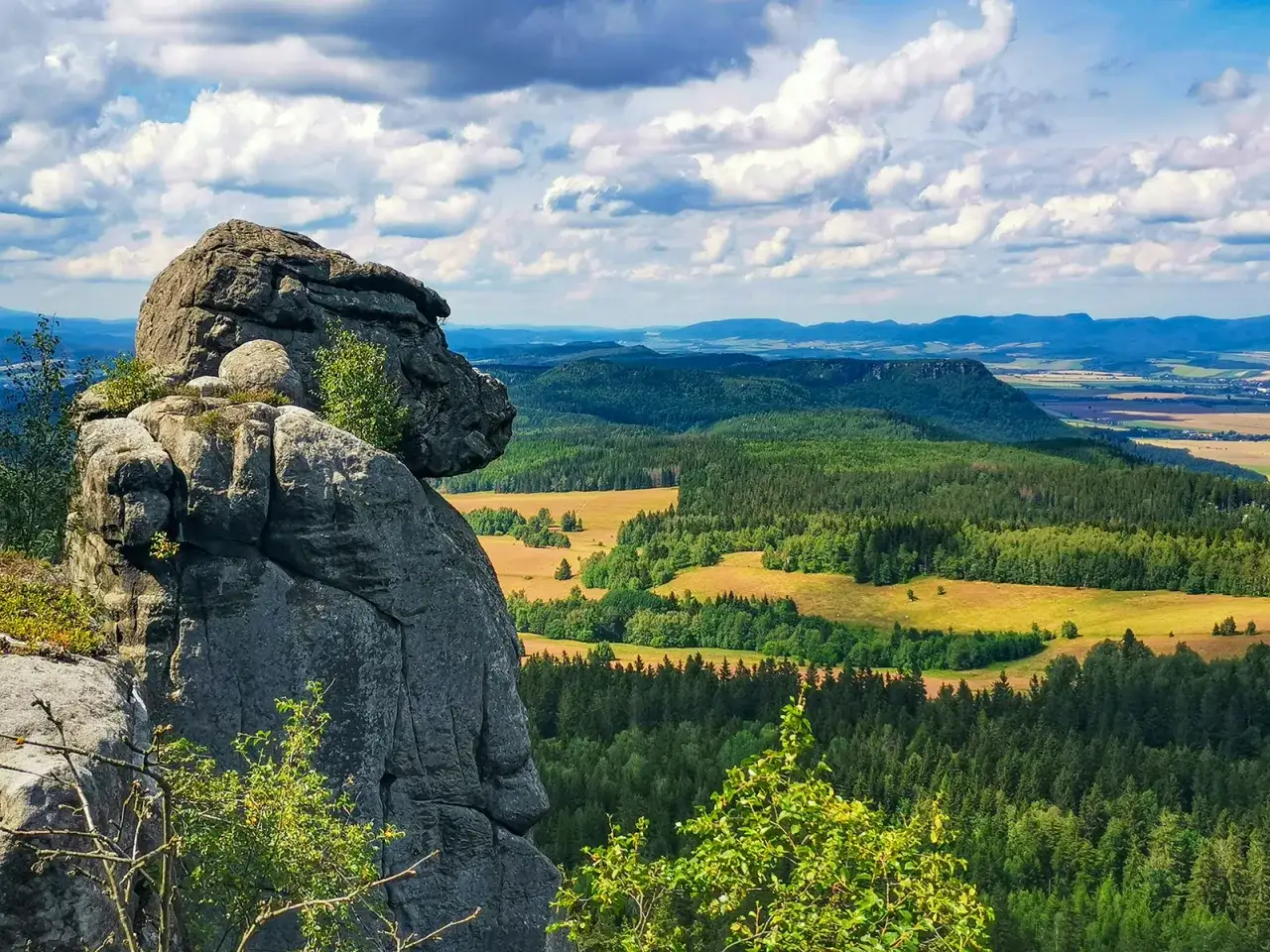 Skalny olbrzym Szczeliniec Wielki góruje nad zielonymi lasami i polami. Chmury na niebie dodają dramatyzmu krajobrazowi.