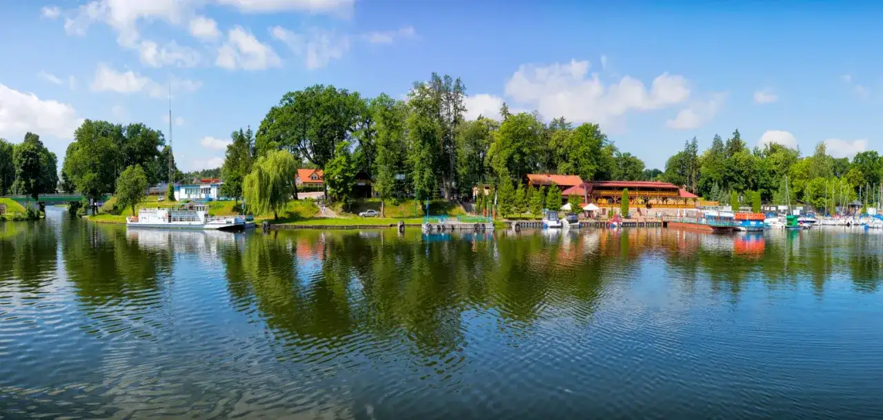 Malowniczy widok na port i jezioro w Rucianem Nidzie. Widać tu cumujące łodzie, restaurację z drewnianą architekturą i zielone drzewa. To idealne miejsce na wypoczynek i podziwianie urok&oacute;w Mazur.