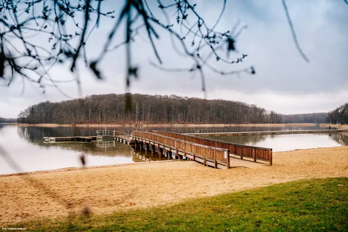 Drewniany pomost i piaszczysta plaża nad jeziorem w Insku, otoczone lasem.