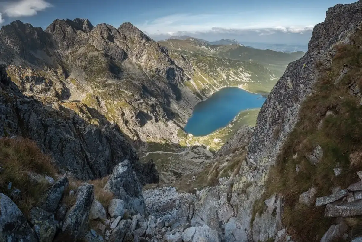 Widok z g&oacute;ry na skaliste zbocze i turkusowe jezioro w dolinie. W oddali majaczą kolejne pasma g&oacute;rskie. Jakby zadni granat.