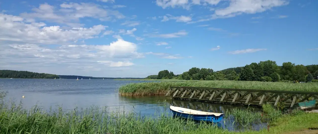 Spokojne jezioro Charzykowskie z ł&oacute;dką przy drewnianym pomoście i żagl&oacute;wkami na horyzoncie.