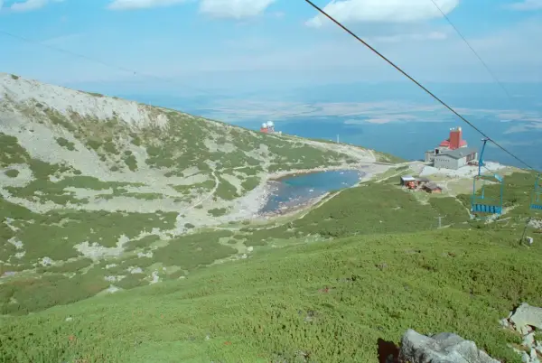 Widok na skaliste pleso otoczone zielenią i kamieniami. W oddali budynek z czerwoną wieżą i kolejka linowa.